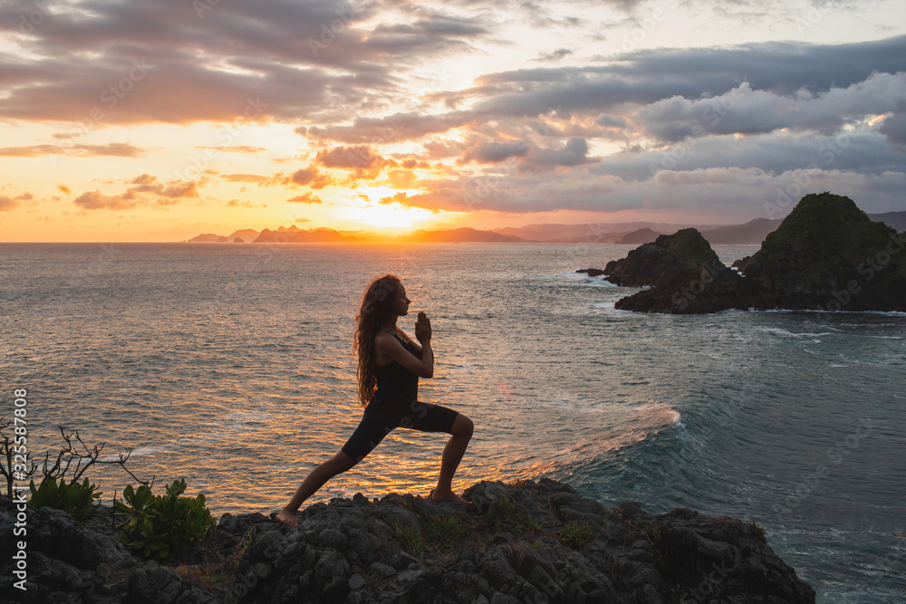 Wall mural young slim woman practicing yoga at sunset with beautiful ocean and mountain view. workout outdoors,