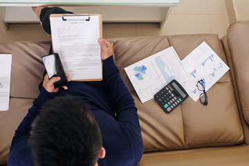 Naklejka premium Businessman sitting on sofa with financial charts and reading partnership agreement, view from above