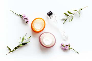 Cosmetologist tools. Dermaroller, creams near leaves and flowers on white background top-down flat lay