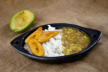 lateral view of an artisanal mud dish containing meat hash soup with rice and slices of fried plantains and an out of focus avocado in the back on a jute mat