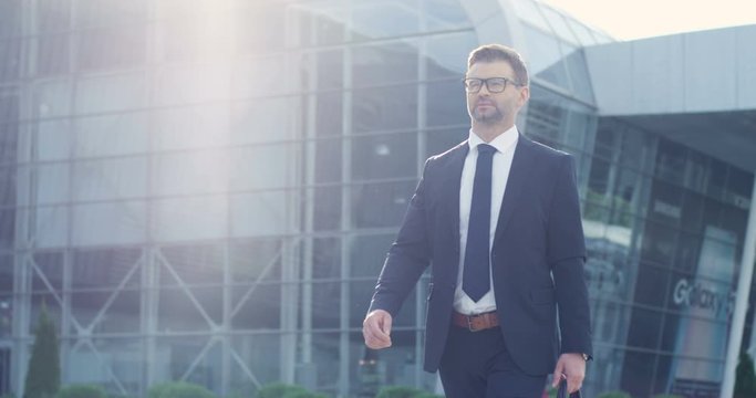 Stylish handsome young confident businessman in suit, tie and glasses walking outdoors in sunlight at aeroport. Caucasian successful man stepping outside big glass urban building on background.