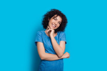 Charming woman with curly black hair is touching her chin and posing on a blue background in a dress