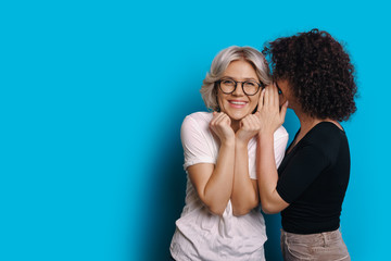 Curly haired caucasian girl is talking something secret to her blonde friend who is wearing eyeglasses and posing on a blue blank space background - obrazy, fototapety, plakaty