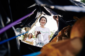 Smiling young Asian volunteers putting garbage that found in park in big sack, view from the sack