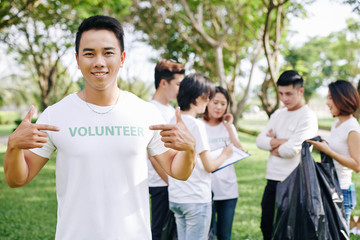 Smiling young Vietnamese man pointing at volunteer inscription on his t-shirt while his team distributing work in background