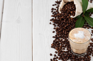 Cappuccino or latte with milk froth in a glass and coffee beans in a bag of coffee and coffee leaves on a white wooden background. Soft focus, top view, copy space for text.