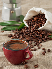 Red cup of coffee (espresso) and coffee beans in burlap sack on a burlap background. Close up, soft focus. Near coffee leaves and coffee pot.