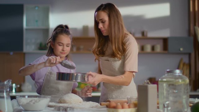Woman teaching daughter to sieve flour on dough at kitchen