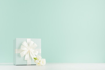 Springtime festive background with white freesia flowers, standing closed square gift box with ribbon on green mint menthe interior on white wood table.