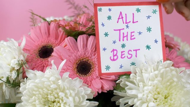 All the best message card with a beautiful bouquet of pink and white flowers. Woman hands placing 'all the best' wishing card with gerbera daisy flowers and chrysanthemums flowers in the background