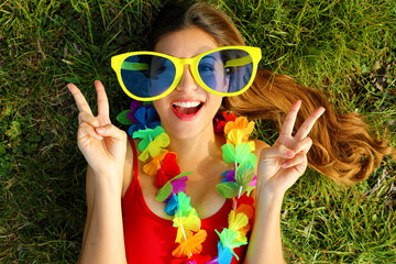 Girl wake up on grass after Carnival party. Young woman with big funny sunglasses and carnival garland lying on grass showing victory V sign with hands.