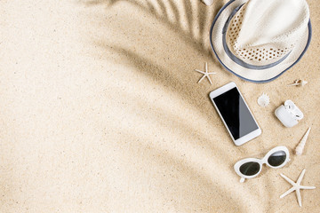 White summer hat, sunglasses, phone, headphone, sea shells and starfish with palm tree branch shadow on sand background, copy space, flat lay