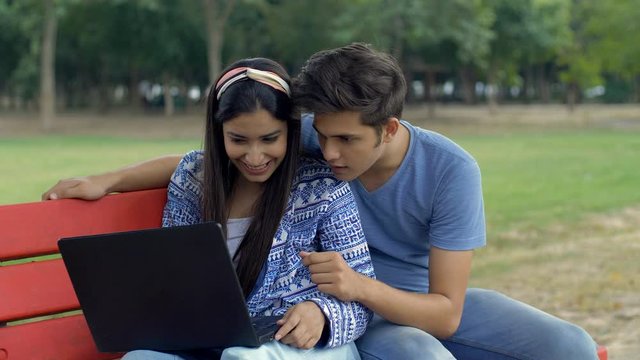 Young college couple having fun while sitting together in the campus - leisure time. Cute Indian boyfriend and girlfriend laughing together while watching funny videos on their laptop during a free...