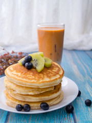 Stack of pancakes with topping, kiwi and blueberry.placed in a white plate on a blue wooden table.Eat with milk tea in the glass.