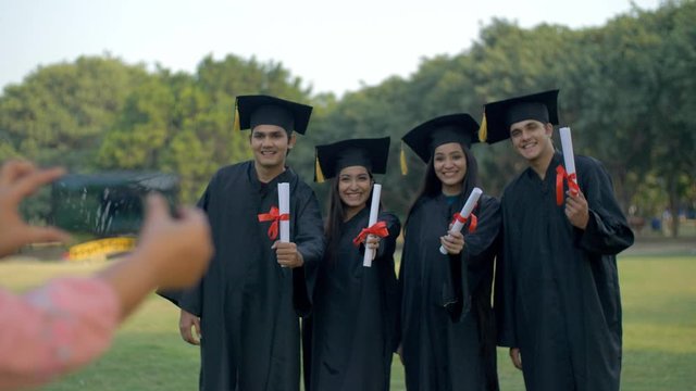 Happy group of young teenagers posing for the photo on convocation ceremony. Pan shot of woman hands clicking pictures of excited young college pupils wearing gowns and caps on their graduation day