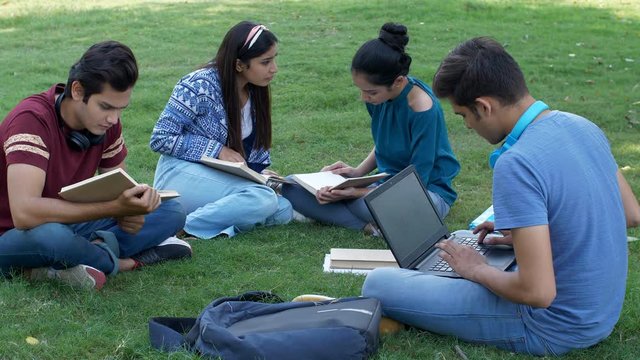Group of four young students studying together in the open-air - college scene. Cute Indian boy typing on his laptop and smiling at the camera while sitting with his friends in the central lawn - l...