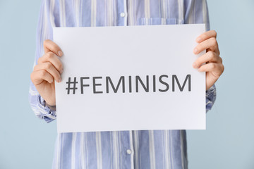 Woman holding paper sheet with word FEMINISM and hashtag on color background, closeup