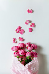 Flower bouquet of pink roses and other mixed flowers wrapped in soft pink paper. The white background is filled with randomly scattered pink petals