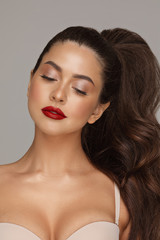 Stock photo portrait of a beautiful young girl with perfect make up and red lips. She has gorgeous thick hair done up in tail. Looking down, wearing bra. Studio shot.