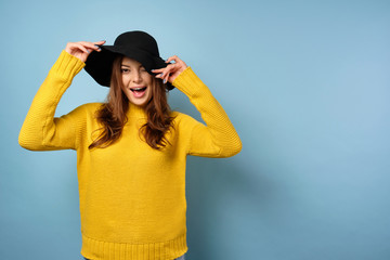A beautiful brunette in a yellow sweater is standing on a blue background and winking, she covers her face with a hat.