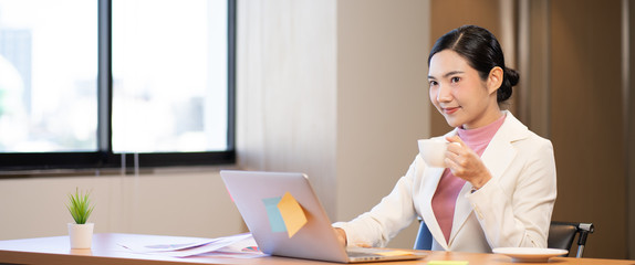 Happy attractive Asian businesswoman working on laptop and holding coffee cup with smile in Modern office or living room with copy space,E-commerce,internet technology, startup small business concept