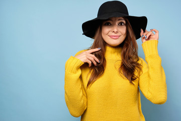 A brunette in a yellow sweater and hat stands on a blue background in surprise with round eyes and holding the edge of the hat