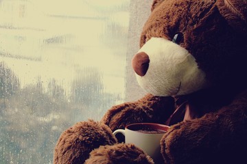 A brown toy bear sits on a window with a cup of tea and looks at raindrops on the glass.