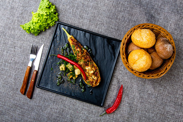 Eggplant baked with cheese and sun-dried tomatoes lies on a black square plate with a long red pepper. A plate on a gray canvas, next to a bread basket with round bread, a bunch of salad