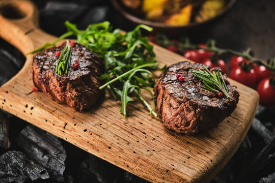 Grilled fillet steaks on wooden cutting board. Succulent thick juicy portions of grilled fillet steak served with tomatoes and roast potatoes on an old wooden board.