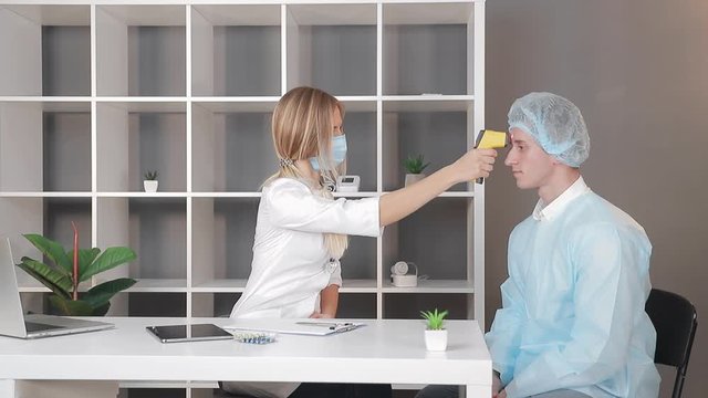 Doctor in a white coat and a medical mask measures the temperature of a patient in the medical office with an infrared non-contact thermometer.