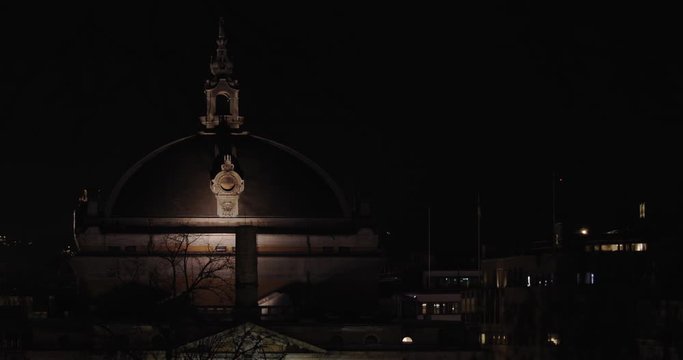 Slow motion wide shot of the dark and illuminated National Theatre building next to Karl Johan street with tree branches moving in front of camera in parallax motion, at night in Oslo Norway.