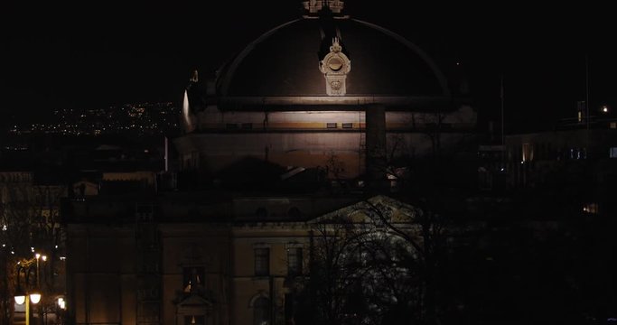 Slow motion wide tilt reveal shot of the dark and illuminated National Theatre building in Karl Johan street with tree branches moving in front of camera in parallax motion, at night in Oslo Norway.
