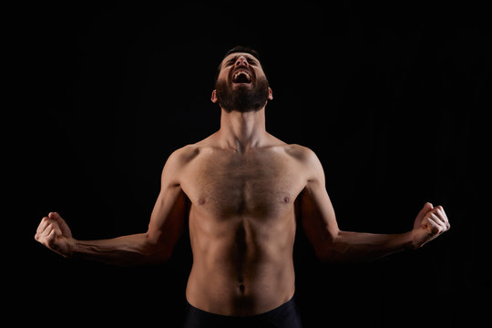Caucasian young man with beard, screaming in rage, no shirt, with open arms, muscular body, on black background, horizontal