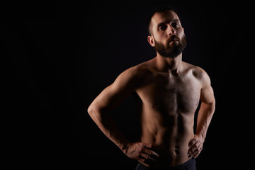 Fototapeta na wymiar Caucasian young man with beard, serious, shirtless, muscular body, on black background looking straight ahead with hands on waist, horizontal
