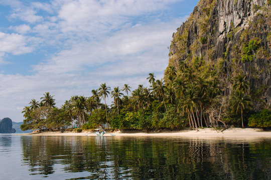 Traditional philippinian boat near idyllic beach with palm trees and mountain. Tropical landscape of Ellis island in Philippines, near Palawan. Amazing nature in Asia. Exotic beach and jungle.  