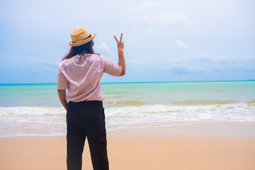 Summer vacation asian girl tourist wearing a hat standing relax and Hold two fingers to show victory. at the sand beach happily. copy space background.