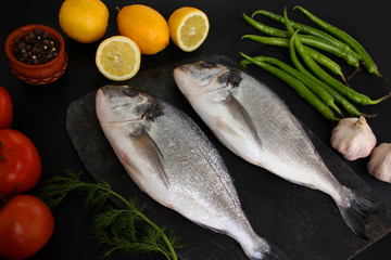 lemons, tomatoes, hot peppers and two carcasses fresh raw atlantic saltwater fish Sparus aurata with shiny silver scales lie on a black background, cooking concept