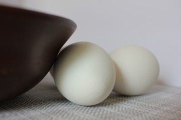 White chicken eggs near brown ceramic bowl on a white texture background