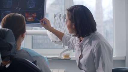 A female dentist shows an MRI image on the screen and an x-ray of the patient's teeth. Discussion of the treatment plan and healthy smile. Recovery after an accident - Powered by Adobe