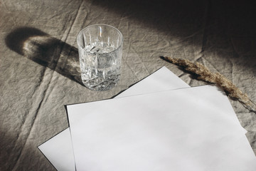 Stationery still life scene. Glass of water, dry grass on beige linen table cloth background in sunlight. Blank paper sheets, cards mockup scene with long harsh shadows. High angle view.