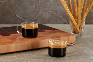 Two shots of espresso with thick foam in clear glass mugs with handles, standing at the cutting Board against the background of a bowl of Italian cookies.