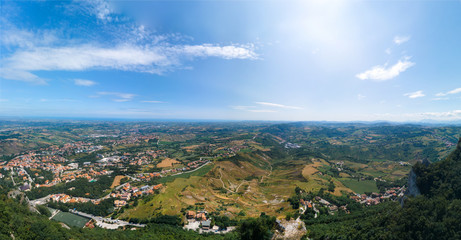 Fototapeta premium High top aerial view Italy landscape in summer, hills and farms, fields and forests, in the far mountains. panoramic wallpaper. In the distant city of Rimini and the sea. San Marino