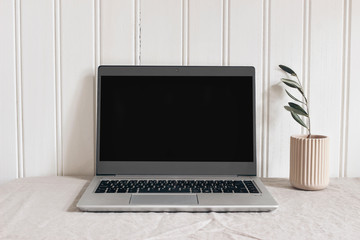 Feminine modern sparse workspace composition. Notebook mock-up scene. Pink ceramic vase with olive branch and laptop on beige linen tablecloth background. White wooden wall. Scandi design.