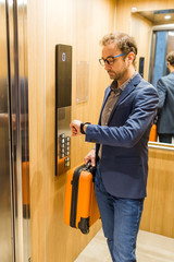 Business traveler holding baggage and checking time on his watch while standing in elevator.