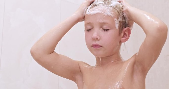 Little boy soaps hair with shampoo in the shower. Child  washes under stream of warm water. Caucasian kid takes shower in the bathroom.  European boy rinses hair with water. Slow motion.