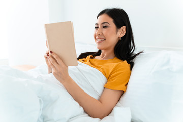 Woman lies in bed indoors at home reading book.