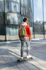 Young hipster in red jacket, sunglasses, jeans, white sneakers and with green backpack riding on longboard by paving stone. Building on the background. Concept of leisure activity, urban and sport.