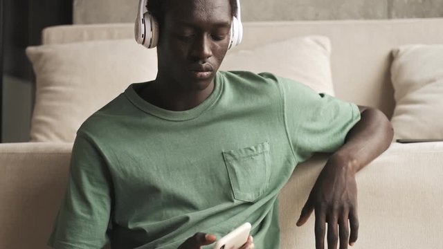 A calm young african american man with wireless headphones is listening to the music while using his smartphone sitting in the living room