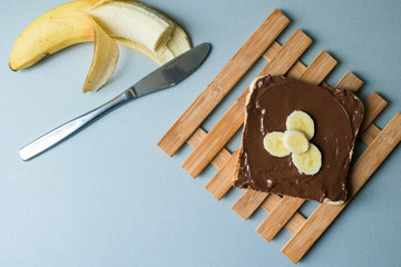A delicious piece of toast with chocolate spread and banana slices on a modern kitchen countertop with a kitchen knife, plate and vintage wooden surface