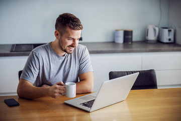 Young smiling man dressed casual sitting at dining table, holding mug with morning coffee and...